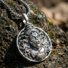 Antique silver pendant with intricate deity face design on a metal chain, placed on a rock