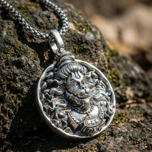 Antique silver pendant with intricate deity face design on a metal chain, placed on a rock